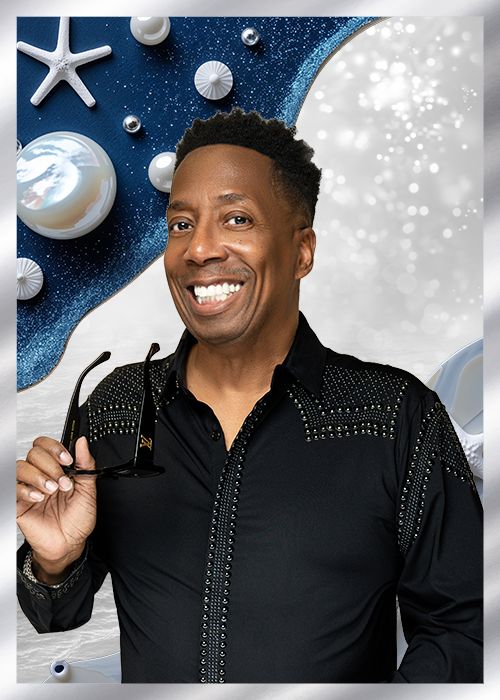 A smiling Black man wearing a black shirt with embroidered designs, standing in front of a blue and white background with starfish and other ocean-themed elements.