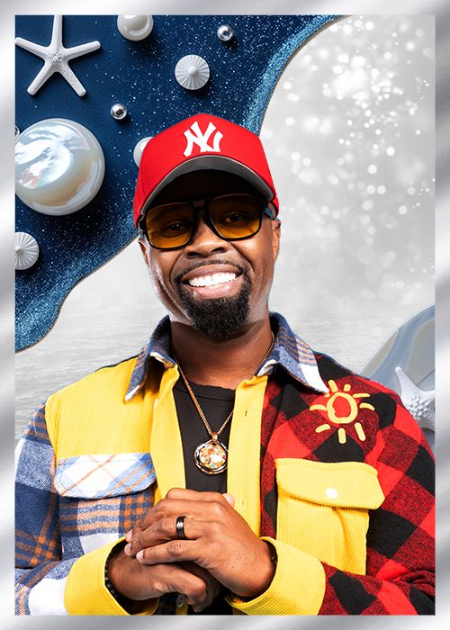 A smiling man wearing a red New York Yankees baseball cap, yellow plaid shirt, and sunglasses stands in front of a blue and white background with starfish and other ocean-themed decorations.