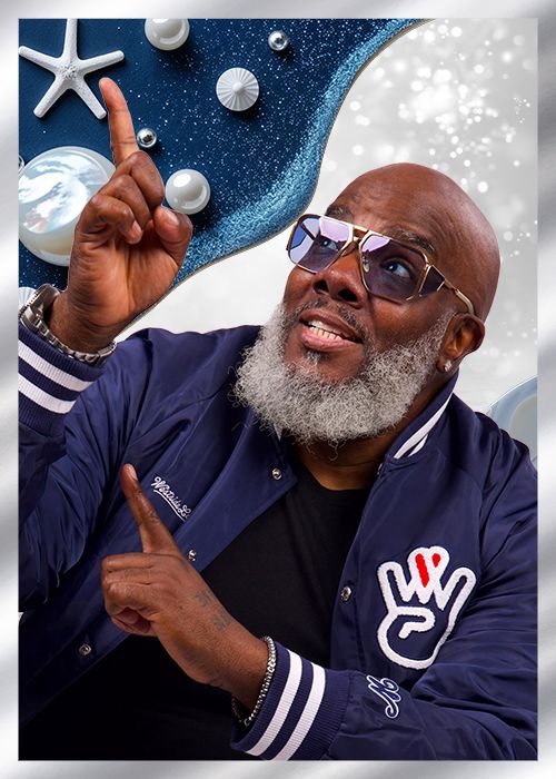 A smiling man with a beard wearing sunglasses and a navy blue jacket with white stripes, pointing upwards against a starry night sky background with various celestial objects.